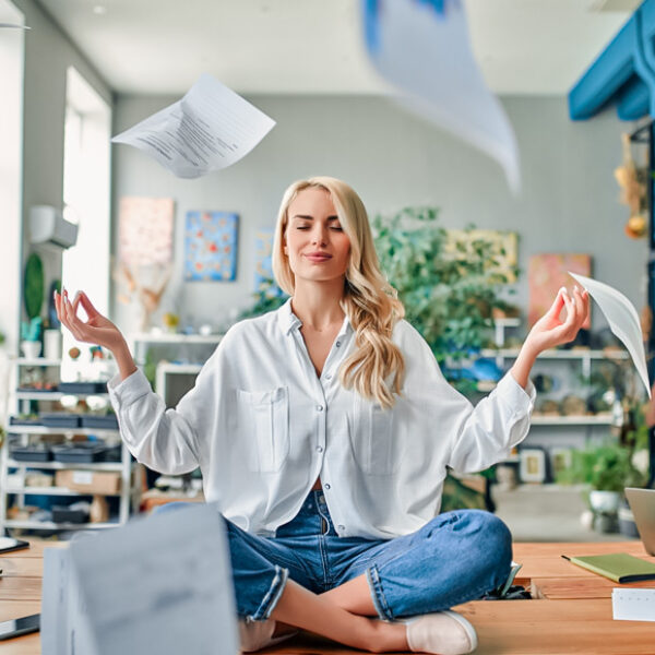 Wellbeing w biurze – kobieta medytująca na biurku w gwarnym biurze typu open space.